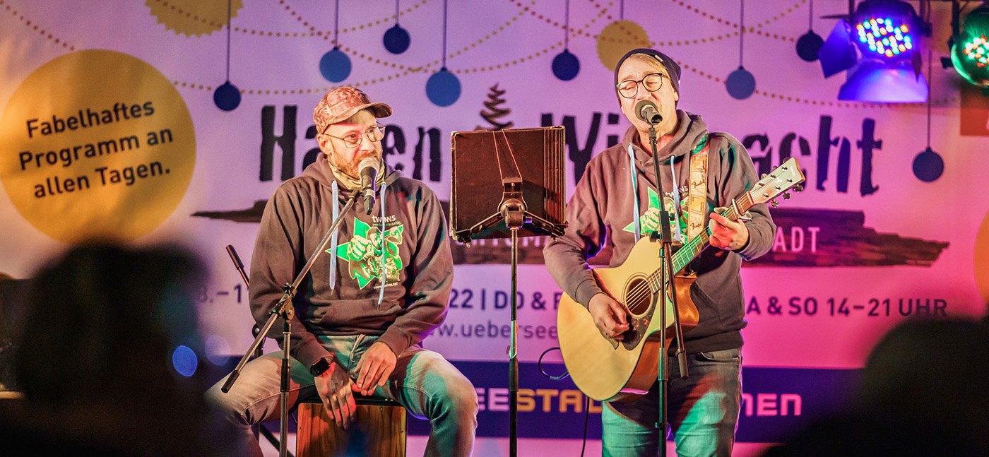Zwei Männer machen Musik auf einer Bühne. Dahinter steht HafenWiehnacht.