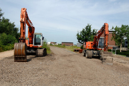 Auf einem Schotterweg stehen zwei Bagger. Im Hintergrund sind wenige Gebäude und Grünflächen erkennbar.