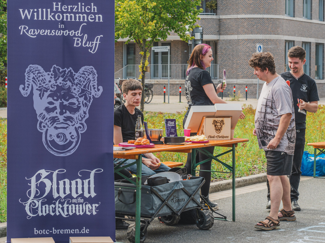 Die Gruppe des Spieletreffs sitzt an einem Infostand und informiert über das Spiel Blood on the Clocktower, dessen lilafarbener Wimpel im Vordergrund flattert Die Gruppe des Spieletreffs sitzt an einem Infostand und informiert über das Spiel Blood on the Clocktower, dessen lilafarbener Wimpel im Vordergrund flattert