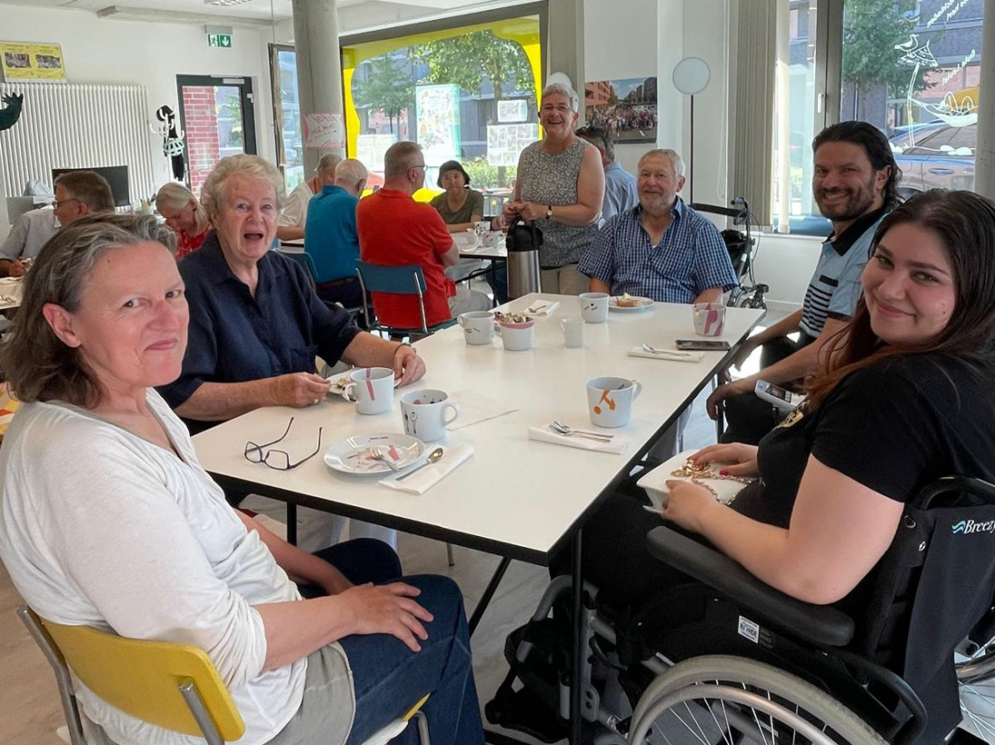 Frauen und Männer sitzen um einen gedeckten Kaffeetisch herum und lächeln in die Kamera, eine der Damen sitzt im Rollstuhl Frauen und Männer sitzen um einen gedeckten Kaffeetisch herum und lächeln in die Kamera, eine der Damen sitzt im Rollstuhl