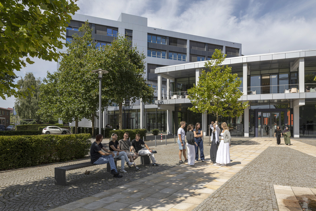 Ein Schulgebäude mit einem Platz, auf dem Bänke stehen. Einige Jugendliche haben sich auf dem Platz versammelt und unterhalten sich. Am Rand steht ein Baum und eine Hecke. Ein Schulgebäude mit einem Platz, auf dem Bänke stehen. Einige Jugendliche haben sich auf dem Platz versammelt und unterhalten sich. Am Rand steht ein Baum und eine Hecke.