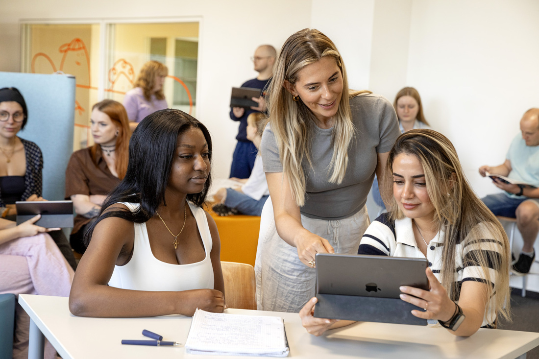 Eine Schulklasse im Unterricht. Eine Lehrerin (stehend) zeigt einer Schülerin (sitzend) etwas auf einem iPad. Um die beiden herum sitzen andere Schüler. Eine Schulklasse im Unterricht. Eine Lehrerin (stehend) zeigt einer Schülerin (sitzend) etwas auf einem iPad. Um die beiden herum sitzen andere Schüler.