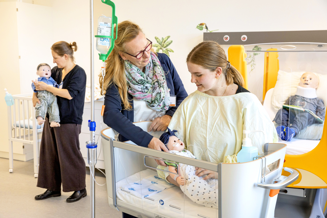 Eine Frau beugt sich im Vordergrund über ein Babybett mit einer Übungspuppe. Im Hintergrund hält eine junge Frau eine weitere Puppe auf dem Arm. Eine Frau beugt sich im Vordergrund über ein Babybett mit einer Übungspuppe. Im Hintergrund hält eine junge Frau eine weitere Puppe auf dem Arm.