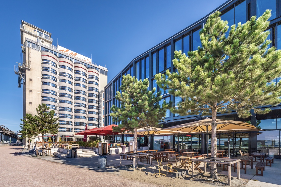 Links ragt das Silo-Hotel John & Will in die Höhe, auf der rechten Seite befindet sich das Reislager mit Sitzgelegenheiten und Bäumen im Vordergrund.