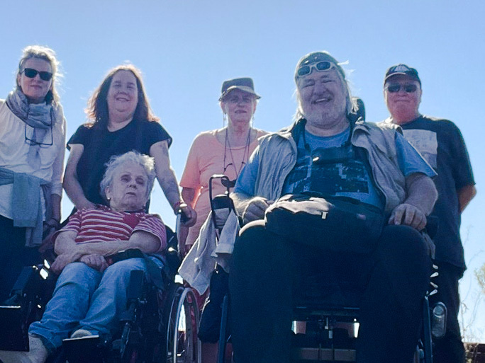 Eine Gruppe von sechs Menschen vor blauem Himmel, die beiden vorderen Personen sitzen im Rollstuhl Eine Gruppe von sechs Menschen vor blauem Himmel, die beiden vorderen Personen sitzen im Rollstuhl